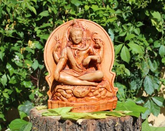 May include: A wooden sculpture of the Hindu deity Shiva, sitting in a meditative pose with his hands in a mudra. He is adorned with a snake around his neck and a third eye on his forehead. The sculpture is painted in shades of brown and orange and has intricate details.