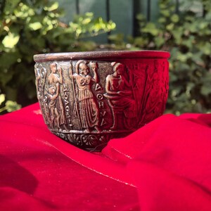Op de afbeelding: Een bronzen beker met een reli&euml;f van figuren en het woord "&Delta;&Omicron;&Eta;&Nu;&Eta;" rond de rand. De beker staat op een rood oppervlak.