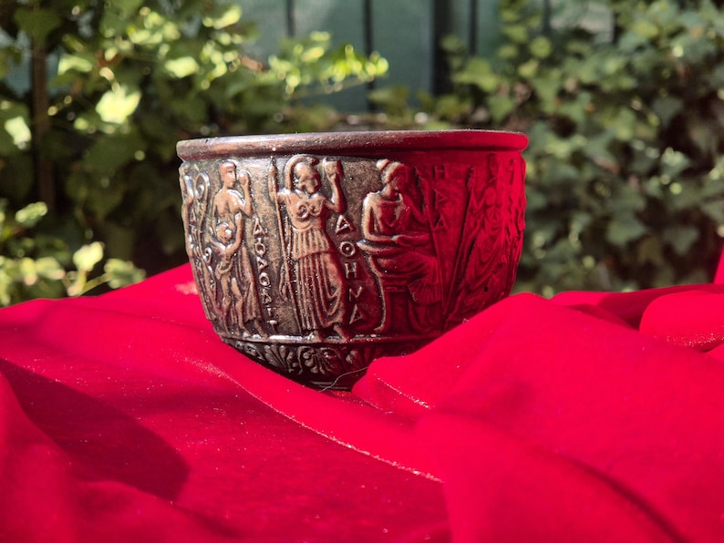 Op de afbeelding: Een bronzen beker met een reli&euml;f van figuren en het woord "&Delta;&Omicron;&Eta;&Nu;&Eta;" rond de rand. De beker staat op een rood oppervlak.