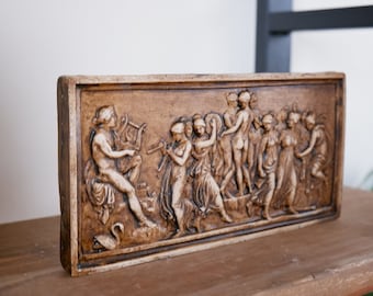May include: A rectangular brown ceramic wall plaque featuring a raised relief sculpture of a procession of figures. The figures are dressed in flowing robes and appear to be playing musical instruments. The plaque has a brown background and a white border.
