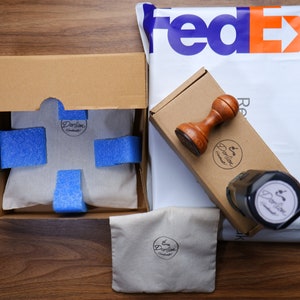 Puede incluir: Una caja de cart&oacute;n marr&oacute;n con un interior blanco y bolitas de embalaje azules. La caja est&aacute; abierta y contiene una bolsa de tela blanca con un logotipo marr&oacute;n que dice "Darlion Handmade". La caja est&aacute; sentada sobre una superficie de madera. Hay una bolsa de env&iacute;o de FedEx en el fondo.