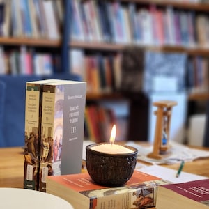 Op de afbeelding: Drie boeken met de titel "Ilkcag Felsefe Tarihi" in het Turks, gestapeld op een houten tafel. Het bovenste boek is deel IV, het middelste boek is deel III en het onderste boek is deel II. Een brandende kaars in een donkerbruine houder staat op de boeken. Op de achtergrond is een wazig boekenrek te zien.