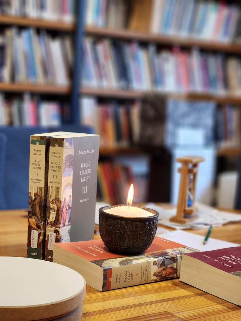 Op de afbeelding: Drie boeken met de titel "Ilkcag Felsefe Tarihi" in het Turks, gestapeld op een houten tafel. Het bovenste boek is deel IV, het middelste boek is deel III en het onderste boek is deel II. Een brandende kaars in een donkerbruine houder staat op de boeken. Op de achtergrond is een wazig boekenrek te zien.