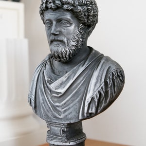 Puede incluir: Un busto de escultura gris de un hombre con barba y cabello rizado. El busto est&aacute; sobre un pedestal con la inscripci&oacute;n "M. Aurelius" en la parte delantera.