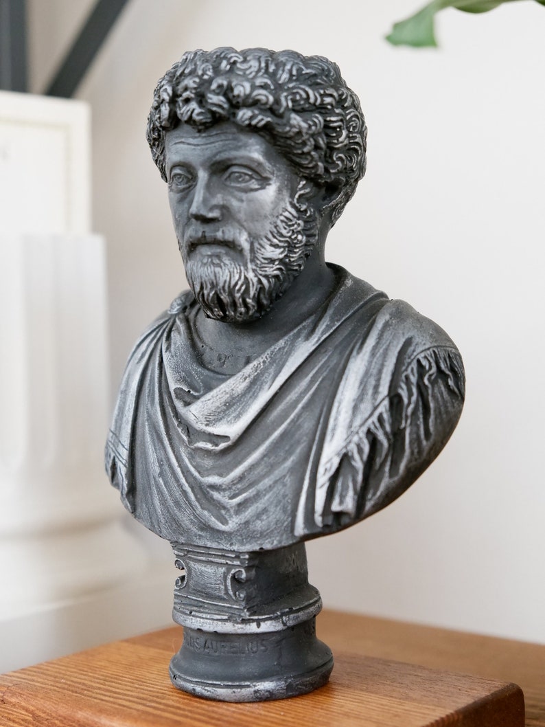 Puede incluir: Un busto de escultura gris de un hombre con barba y cabello rizado. El busto est&aacute; sobre un pedestal con la inscripci&oacute;n "M. Aurelius" en la parte delantera.