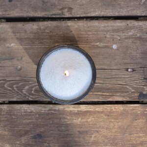 Op de afbeelding: Een witte kaars in een zwarte houder is aangestoken en brandt. De kaars staat op een houten oppervlak.