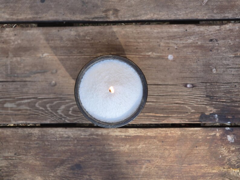 Op de afbeelding: Een witte kaars in een zwarte houder is aangestoken en brandt. De kaars staat op een houten oppervlak.
