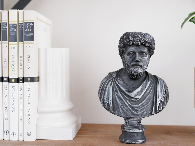 Puede incluir: Una escultura de busto gris de un hombre con barba y pelo corto. El busto est&aacute; sobre un pedestal negro con las palabras "MAGOS AUREL" inscritas en la parte delantera. El busto est&aacute; sentado en un estante de madera con un separador de libros blanco y un conjunto de libros detr&aacute;s.