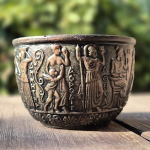 Op de afbeelding: Een kleine, ronde, bronzen beker met een decoratief ontwerp van figuren en Griekse letters. De beker staat op een houten oppervlak.