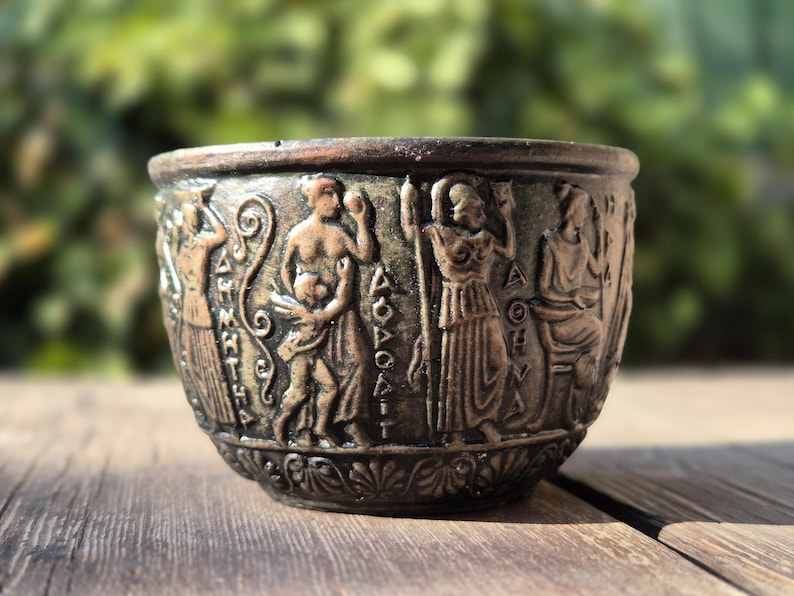 Op de afbeelding: Een kleine, ronde, bronzen beker met een decoratief ontwerp van figuren en Griekse letters. De beker staat op een houten oppervlak.