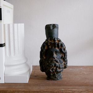 Puede incluir: Un busto esculpido en negro y gris de un hombre con barba y pelo largo. El busto está sentado sobre una superficie de madera delante de un sujetalibros blanco.