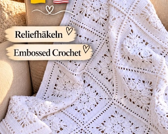 Puede incluir: Una manta de ganchillo blanca con motivos cuadrados que presentan intrincados patrones florales y texturas en relieve. El texto en la imagen dice: Anleitung Häkeldecke, Reliefhäkeln y Embossed Crochet, junto a iconos de banderas alemana y estadounidense.