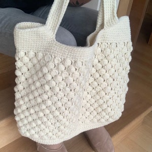 May include: A white crocheted tote bag with a textured, bubble-like design. The bag has two long straps.