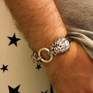 K&ouml;nnte beinhalten: Ein silbernes Armband mit Totenkopf- und Federdesign. Das Armband ist aus Leder und hat einen silbernen Verschluss.