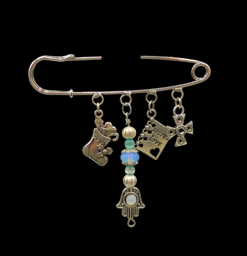 May include: Silver safety pin with charms including a stocking, a hamsa hand, a cross, and a tag that says "Secret Love".