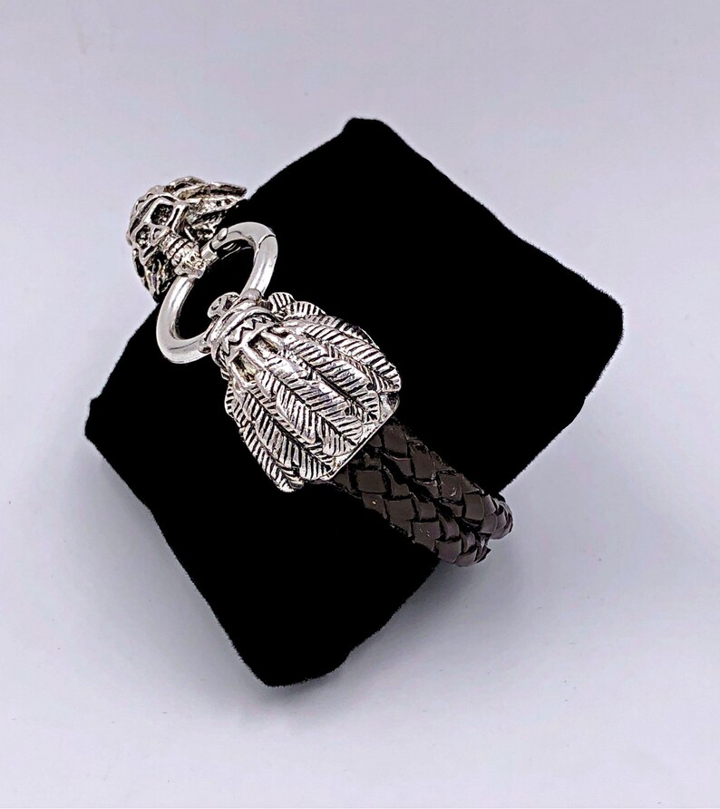 K&ouml;nnte beinhalten: Ein braunes Lederarmband mit einem silbernen Totenkopf- und Feder-Verschluss.