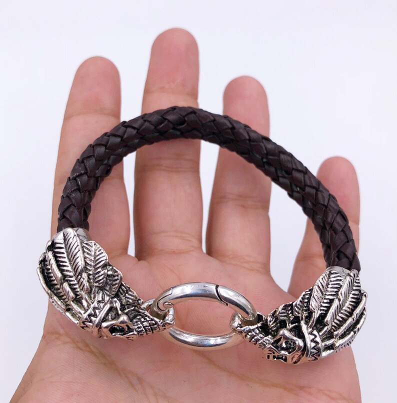 K&ouml;nnte beinhalten: Ein braunes geflochtenes Lederarmband mit silbernen Drachenkopf-Akzenten und einem silbernen Verschluss.