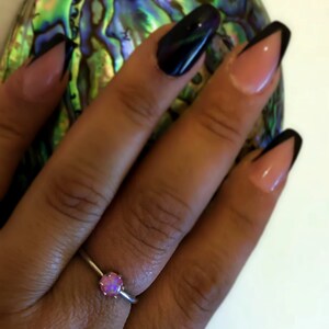 May include: A silver ring with a pink opal gemstone. The ring is on a person's finger. The person's nails are painted black and pink.
