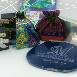 May include: A variety of packaging options for jewelry, including a black box with a white ribbon, a small pouch with a colorful pattern, a clear bag with a gold design, a holographic box, and a blue agate slice with the text "MorMalas - Jewelry Design -" engraved on it.