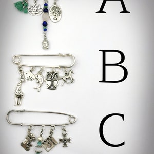 May include: Three silver safety pin brooches with dangling charms. The first brooch has a green tassel, a lotus flower, a pink stone, a tree charm, and a Buddha charm. The second brooch has a giraffe, a dolphin, an owl, and a horse charm. The third brooch has a stocking, a heart charm with the text "Secret Love", a pearl bead, and a cross charm.