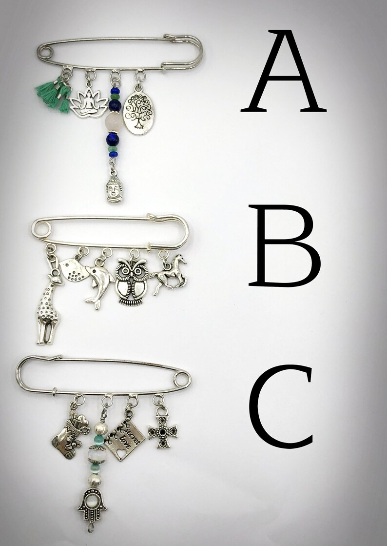 May include: Three silver safety pin brooches with dangling charms. The first brooch has a green tassel, a lotus flower, a pink stone, a tree charm, and a Buddha charm. The second brooch has a giraffe, a dolphin, an owl, and a horse charm. The third brooch has a stocking, a heart charm with the text "Secret Love", a pearl bead, and a cross charm.