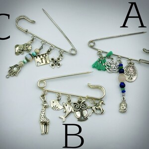 May include: Three silver safety pin brooches with various charms. The first brooch has green tassels, a lotus flower, a tree charm, a pink stone bead, and blue beads. The second brooch has a giraffe, a bird, a dolphin, an owl, and a horse charm. The third brooch has a hand charm, a flower charm, a heart charm with the text "Secret Love", a cross charm, and blue and white beads.