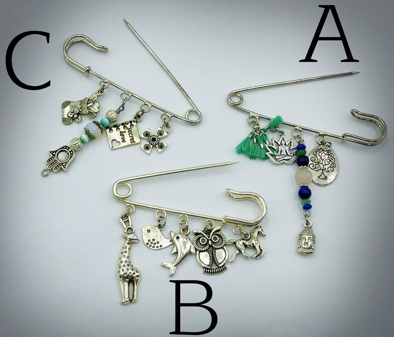May include: Three silver safety pin brooches with various charms. The first brooch has green tassels, a lotus flower, a tree charm, a pink stone bead, and blue beads. The second brooch has a giraffe, a bird, a dolphin, an owl, and a horse charm. The third brooch has a hand charm, a flower charm, a heart charm with the text "Secret Love", a cross charm, and blue and white beads.