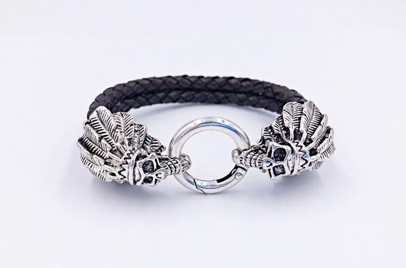 K&ouml;nnte beinhalten: Ein schwarzes Lederarmband mit silbernen Totenkopf- und Federn-Akzenten. Das Armband hat einen runden Verschluss.