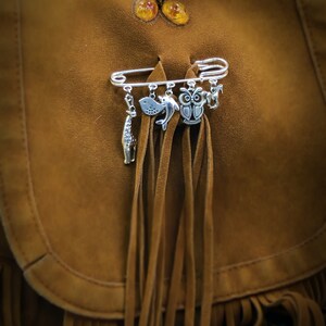 May include: A silver safety pin with multiple charms attached, including an owl, a fish, and a dolphin. The pin is attached to a brown suede bag with fringe.