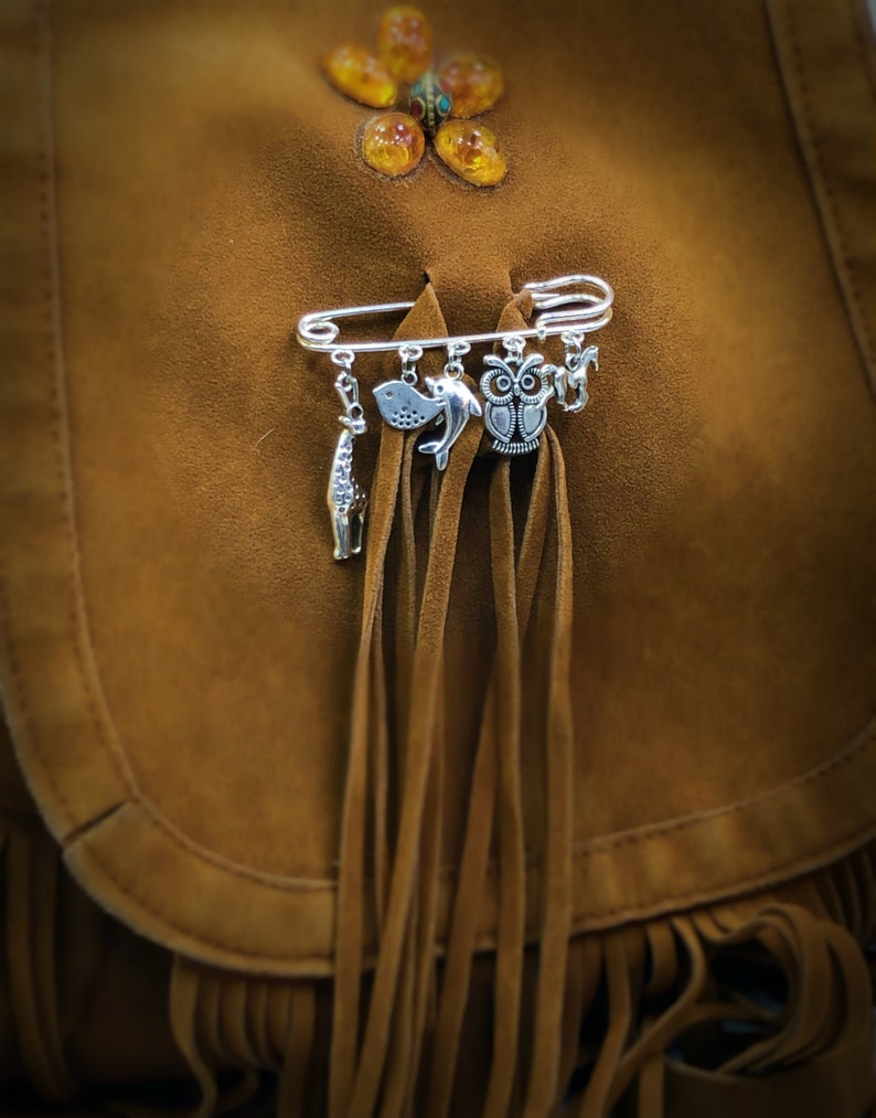 May include: A silver safety pin with multiple charms attached, including an owl, a fish, and a dolphin. The pin is attached to a brown suede bag with fringe.