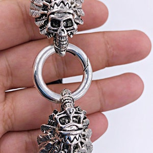 K&ouml;nnte beinhalten: Ein silberfarbenes Metallarmband mit zwei Sch&auml;del-Anh&auml;ngern. Jeder Anh&auml;nger zeigt einen Sch&auml;del, der eine Kopfbedeckung mit Federn tr&auml;gt.