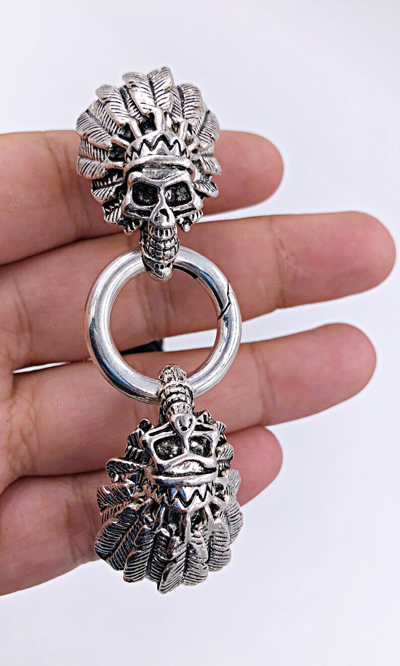 K&ouml;nnte beinhalten: Ein silberfarbenes Metallarmband mit zwei Sch&auml;del-Anh&auml;ngern. Jeder Anh&auml;nger zeigt einen Sch&auml;del, der eine Kopfbedeckung mit Federn tr&auml;gt.