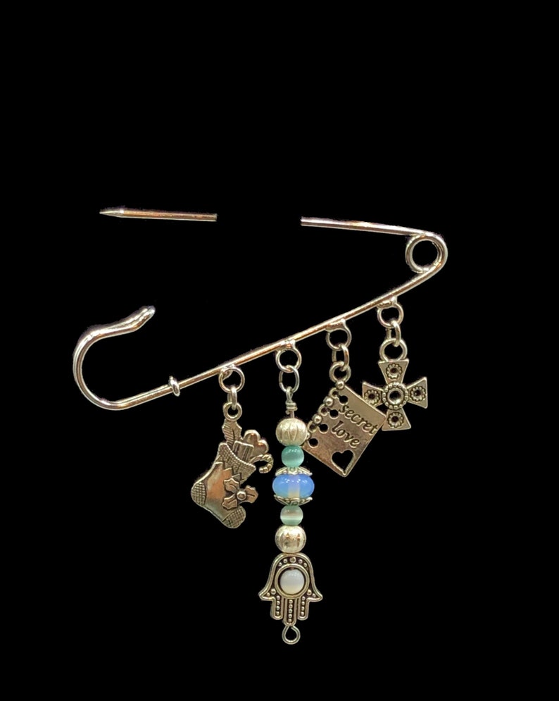 May include: A silver safety pin with charms hanging from it. The charms include a stocking, a cross, a heart with the words "secret love" on it, and a hamsa hand with a blue and white stone.