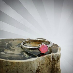 May include: A silver ring with a pink opal gemstone set in a prong setting. The ring is sitting on a piece of petrified wood.