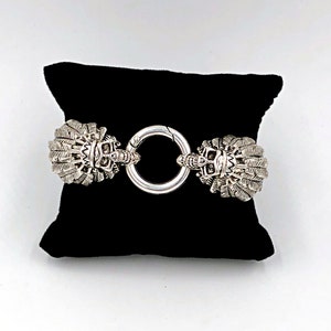 K&ouml;nnte beinhalten: Ein silbernes Armband mit zwei verzierten, detaillierten Sch&auml;delk&ouml;pfen mit Federn an beiden Enden, die durch einen silbernen Ring verbunden sind.