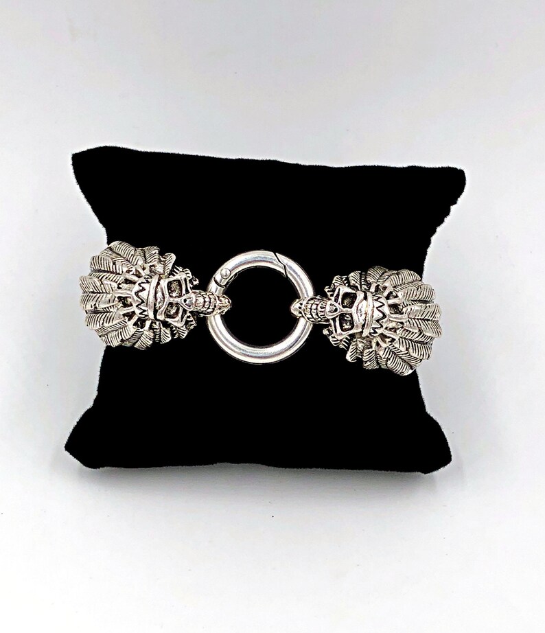 K&ouml;nnte beinhalten: Ein silbernes Armband mit zwei verzierten, detaillierten Sch&auml;delk&ouml;pfen mit Federn an beiden Enden, die durch einen silbernen Ring verbunden sind.