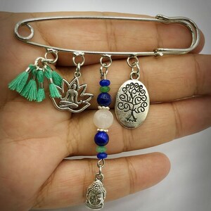 May include: Silver safety pin with charms including a meditating figure in a lotus, a tree of life, a Buddha head, and green tassels. The charms are strung with blue and green beads.