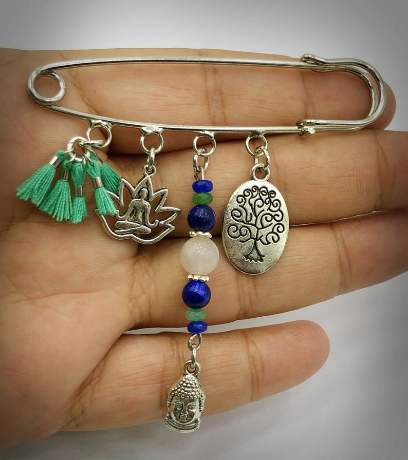 May include: Silver safety pin with charms including a meditating figure in a lotus, a tree of life, a Buddha head, and green tassels. The charms are strung with blue and green beads.