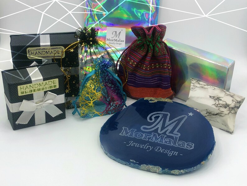 K&ouml;nnte beinhalten: Eine blaue Achatplatte mit dem eingravierten Text "MorMalas - Jewelry Design - ". Die Achatplatte ist von verschiedenen Verpackungsmaterialien umgeben, darunter eine kleine schwarze Schachtel mit einem wei&szlig;en Band, eine kleine wei&szlig;e Schachtel mit einem wei&szlig;en Band, ein kleiner lilafarbener Beutel mit Kordelzug und eine kleine transparente Tasche mit einem goldenen Design.