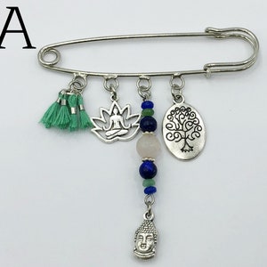 May include: Silver safety pin with charms including a lotus flower, a tree of life, a buddha head, and a tassel. The charms are strung on a chain with blue and green beads.