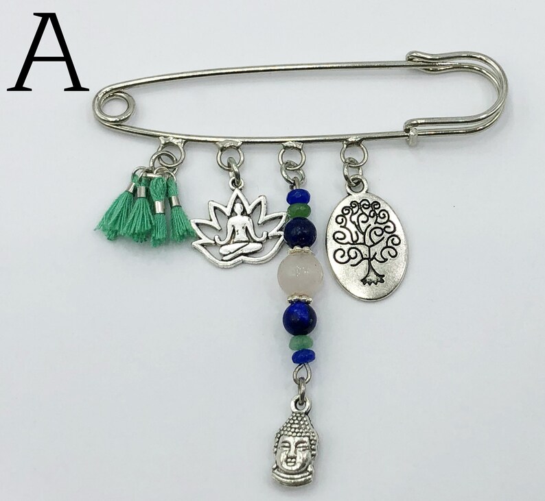May include: Silver safety pin with charms including a lotus flower, a tree of life, a buddha head, and a tassel. The charms are strung on a chain with blue and green beads.