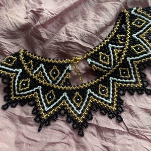 Puede incluir: Un collar de cuentas en negro, dorado y blanco. El collar presenta un patr&oacute;n geom&eacute;trico con un borde festoneado y un cierre dorado. Las cuentas son peque&ntilde;as y est&aacute;n tejidas apretadamente, creando un dise&ntilde;o detallado.