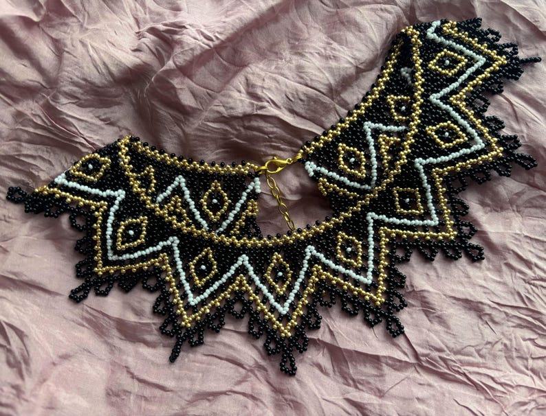 Puede incluir: Un collar de cuentas en negro, dorado y blanco. El collar presenta un patr&oacute;n geom&eacute;trico con un borde festoneado y un cierre dorado. Las cuentas son peque&ntilde;as y est&aacute;n tejidas apretadamente, creando un dise&ntilde;o detallado.