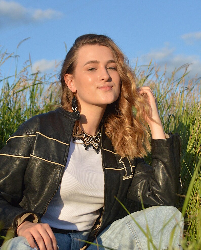 Puede incluir: Chaqueta de cuero negro con rayas color crema y un parche con el n&uacute;mero 1. La chaqueta se lleva sobre una camiseta blanca y combinada con vaqueros azules. La modelo lleva un collar y pendientes de cuentas negras.