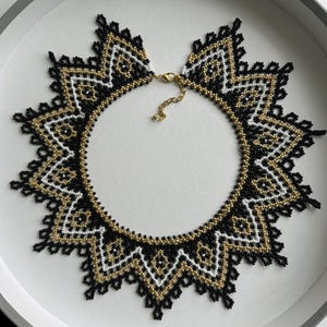 Puede incluir: Un collar de cuentas con un dise&ntilde;o geom&eacute;trico en negro, blanco y dorado. El collar tiene forma circular con un borde decorativo y un cierre dorado.