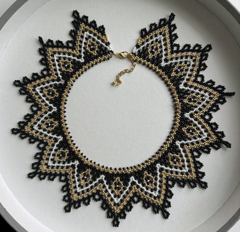 Puede incluir: Un collar de cuentas con un dise&ntilde;o geom&eacute;trico en negro, blanco y dorado. El collar tiene forma circular con un borde decorativo y un cierre dorado.