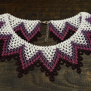 May include: A beaded collar necklace featuring a geometric design. The necklace showcases a pattern of white, purple, and dark burgundy beads. It has a scalloped edge and a silver-tone clasp.