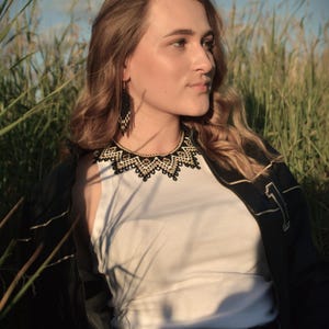 Puede incluir: Una mujer con una camiseta blanca, una chaqueta negra y vaqueros azules, adornada con un collar de cuentas negro y dorado y pendientes a juego. El fondo presenta hierba alta y un cielo azul.