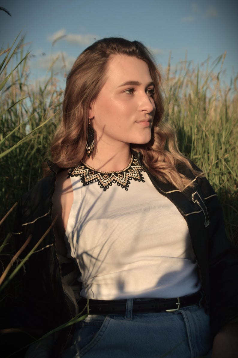 Puede incluir: Una mujer con una camiseta blanca, una chaqueta negra y vaqueros azules, adornada con un collar de cuentas negro y dorado y pendientes a juego. El fondo presenta hierba alta y un cielo azul.