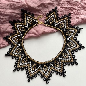 Puede incluir: Un collar de cuentas con un dise&ntilde;o geom&eacute;trico con cuentas negras, doradas y blancas. El collar tiene forma de estrella con un cierre dorado. El intrincado trabajo de cuentas crea un accesorio llamativo.